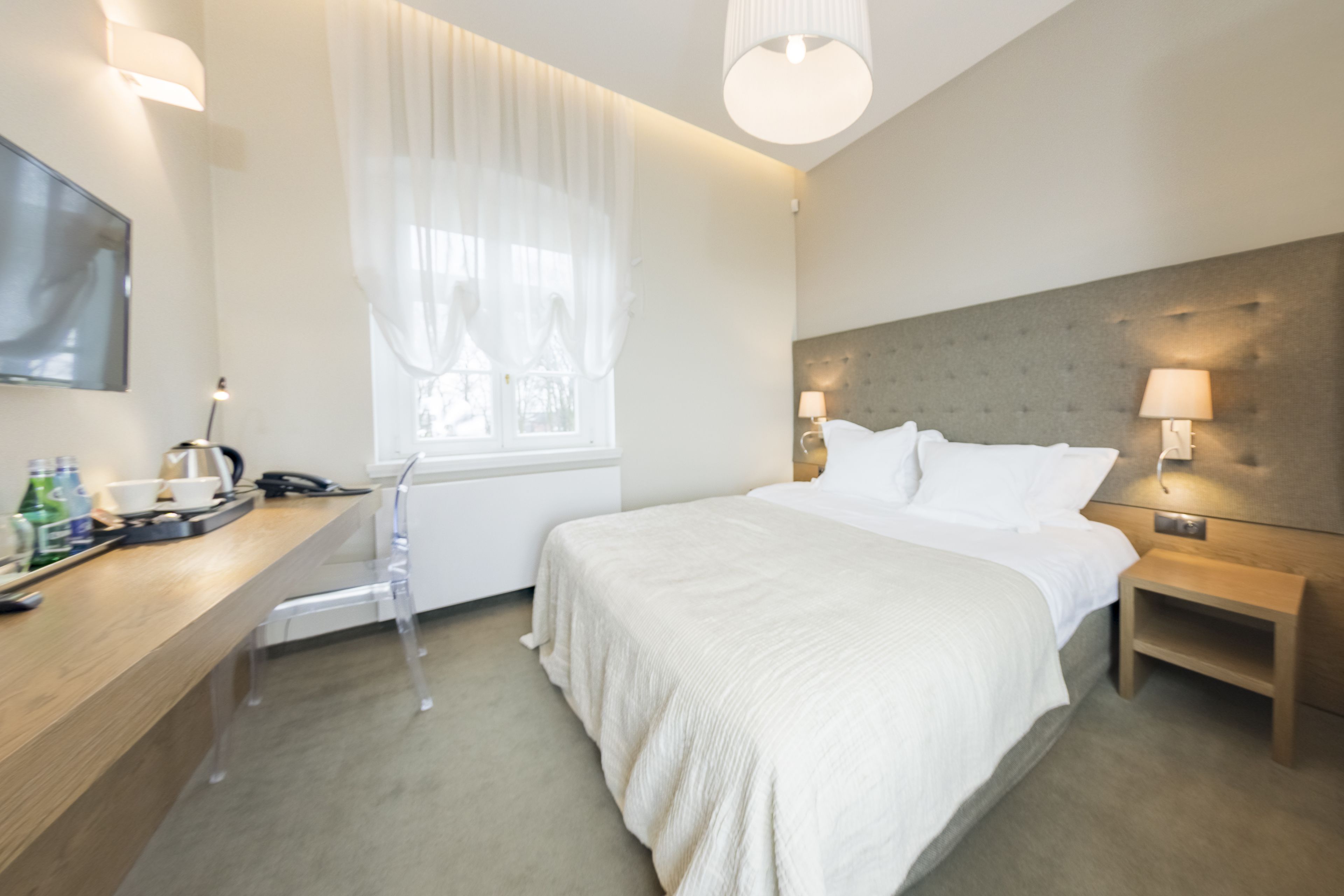 Ein modernes Hotelzimmer mit einem großen Bett, Nachttischlampen, einem Flachbildfernseher, einem Schreibtisch mit Stuhl und einem Fenster mit transparenten Vorhängen.