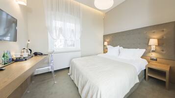 Ein modernes Hotelzimmer mit einem großen Bett, Nachttischlampen, einem Flachbildfernseher, einem Schreibtisch mit Stuhl und einem Fenster mit transparenten Vorhängen.