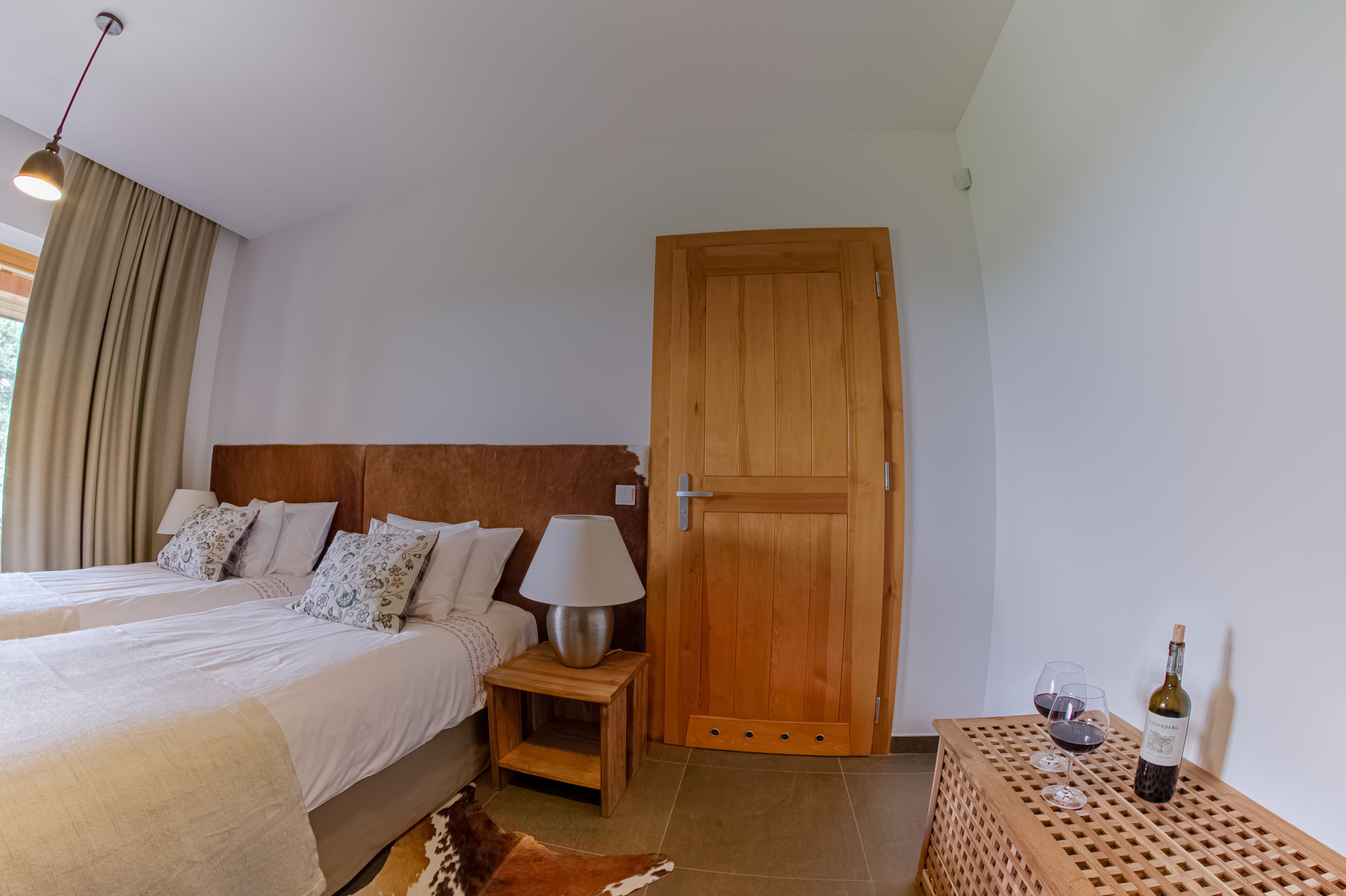A bedroom with two beds, a wooden door, a small table with a bottle and glasses, and a lamp on a nightstand.