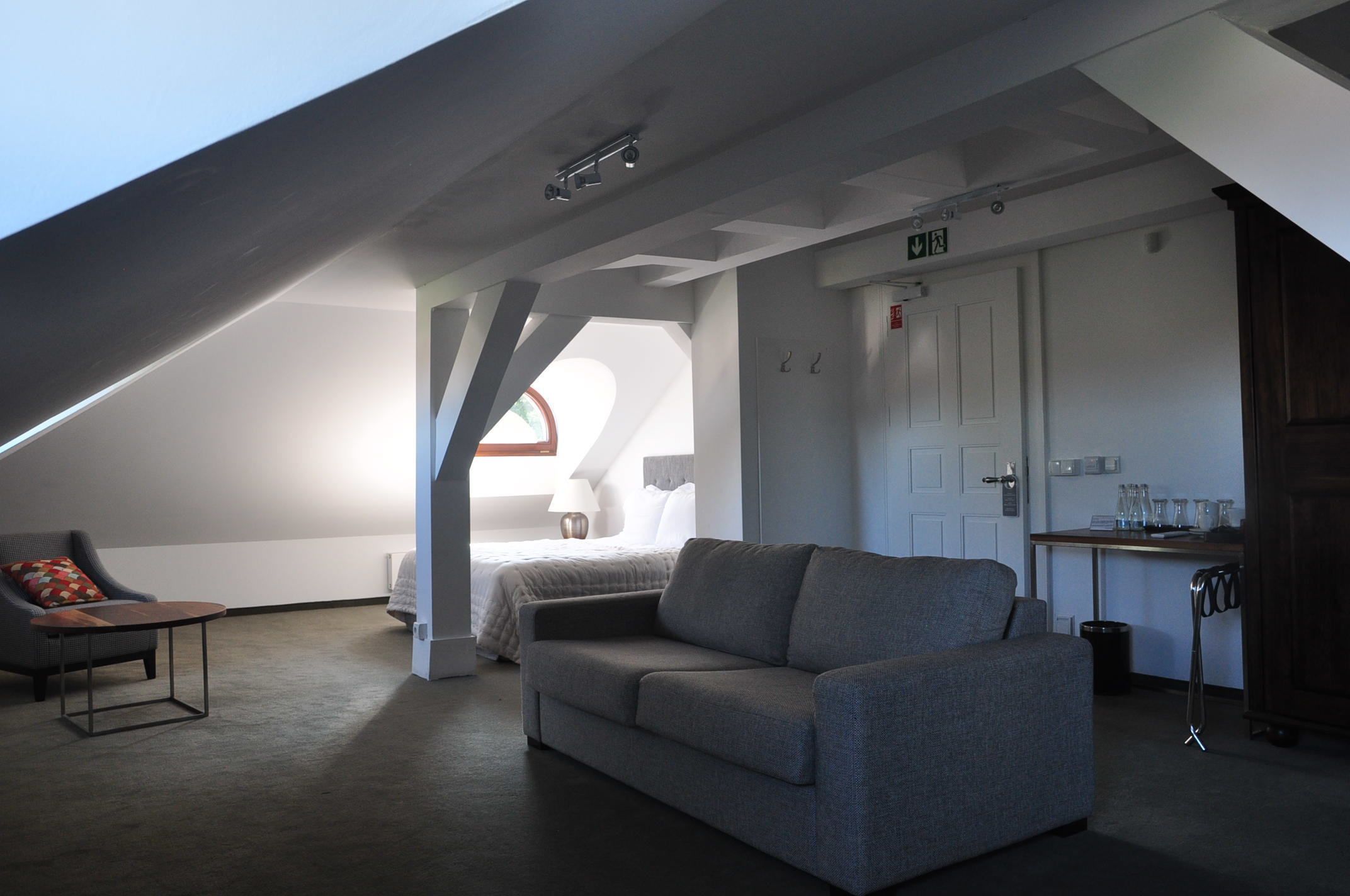 Een moderne loft slaapkamer met een schuine hemel, een grijze bank, een klein tafeltje en een bed met een wit hoofdeinde.
