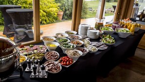 A buffet table with various dishes and drinks, a large metal pot, and a view of a garden through large windows. - Jakubus (Wilków)