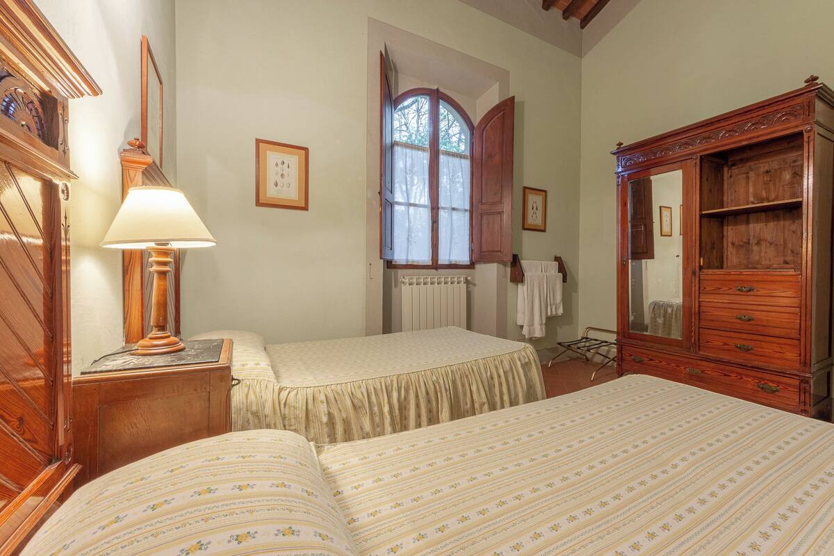 Quarto com duas camas, cômoda de madeira, luminária, janela com persianas e radiador.