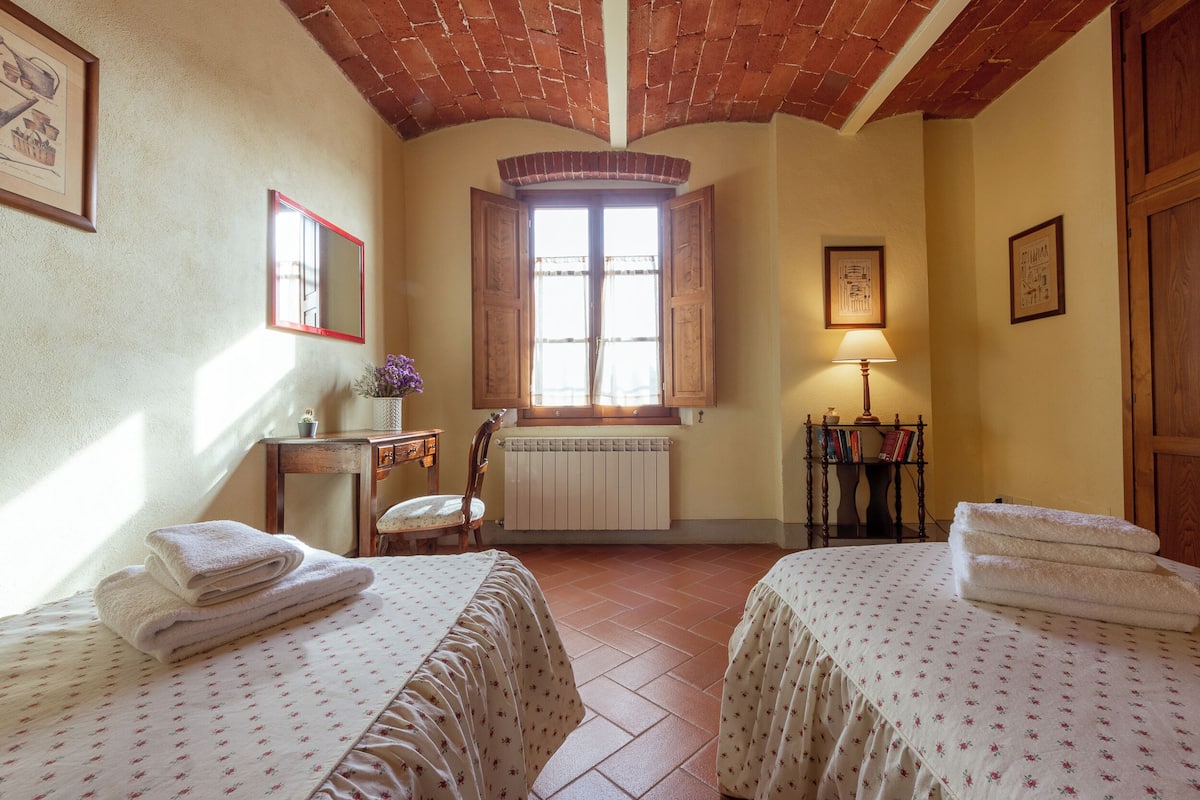 Quarto com duas camas, janela com persianas de madeira, radiador, escrivaninha com abajur e quadros nas paredes.