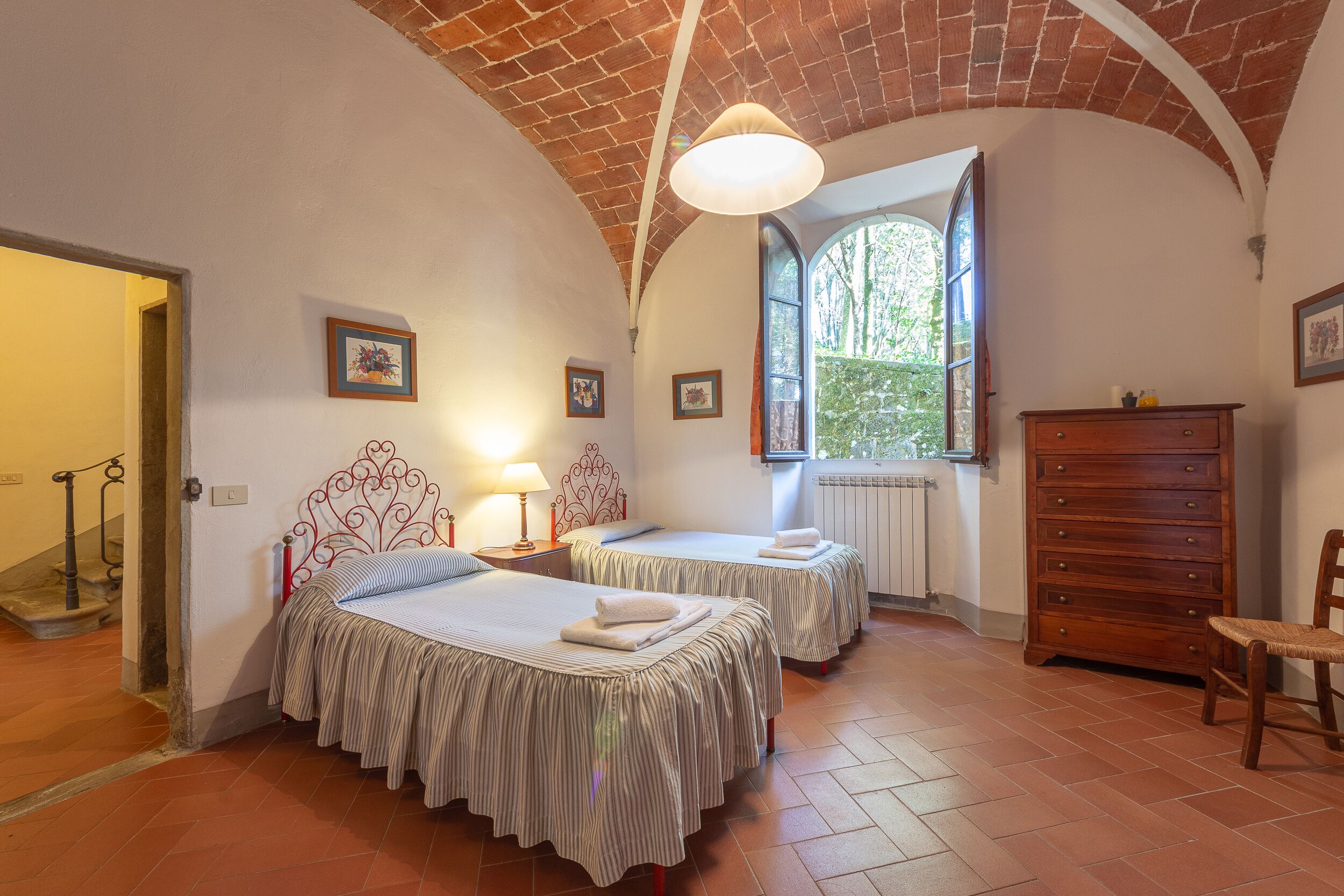 Ein Zimmer mit zwei Betten, einem hölzernen Schminktisch, einem Stuhl und einem Fenster mit Blick auf Bäume.