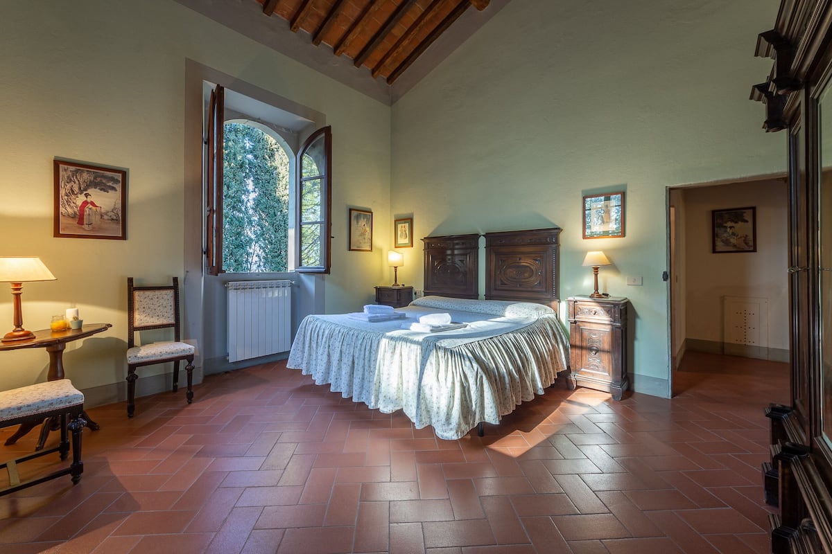 Quarto com uma cama grande, uma cadeira, uma mesa com abajur e uma janela com vista para árvores.