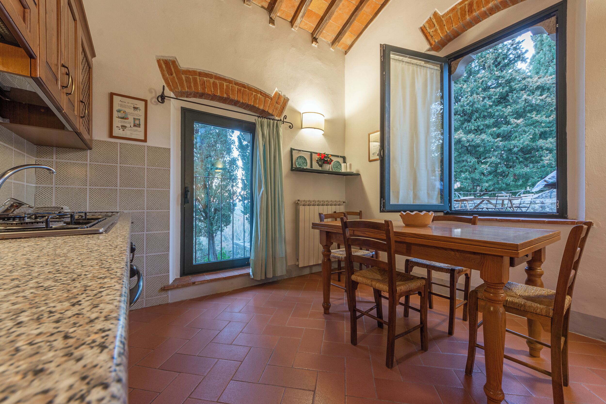 A kitchen with a granite countertop, wooden cabinets, a dining table with chairs, a window with a view of trees, and a brick ceiling.