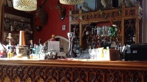 A bar area with wooden paneling, hanging lamps, and a variety of bottles and glasses on shelves. - Chalk And Cheese (King's Lynn)