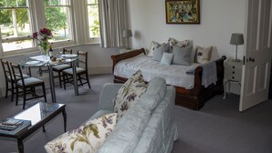 Un dormitorio con cama doble, una zona de estar con sofá y sillones, una mesa de comedor con sillas y una ventana con cortinas.