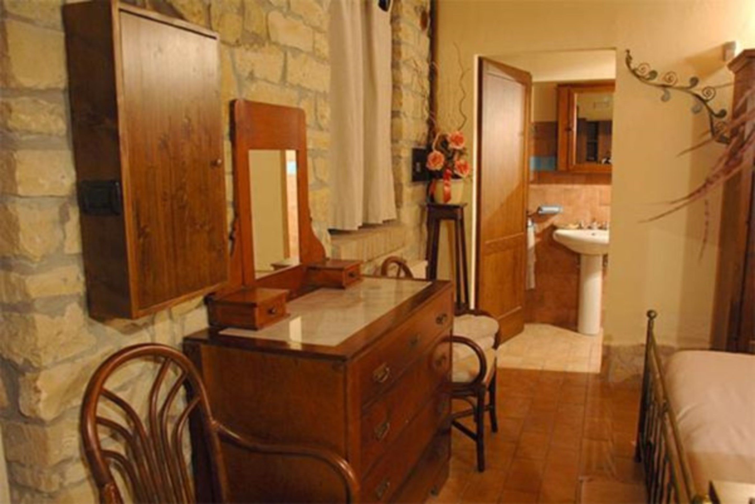 Une chambre avec une commode en bois, une chaise, un lit, un mur en pierre, une salle de bain avec un lavabo et un miroir, et une plante décorative.
