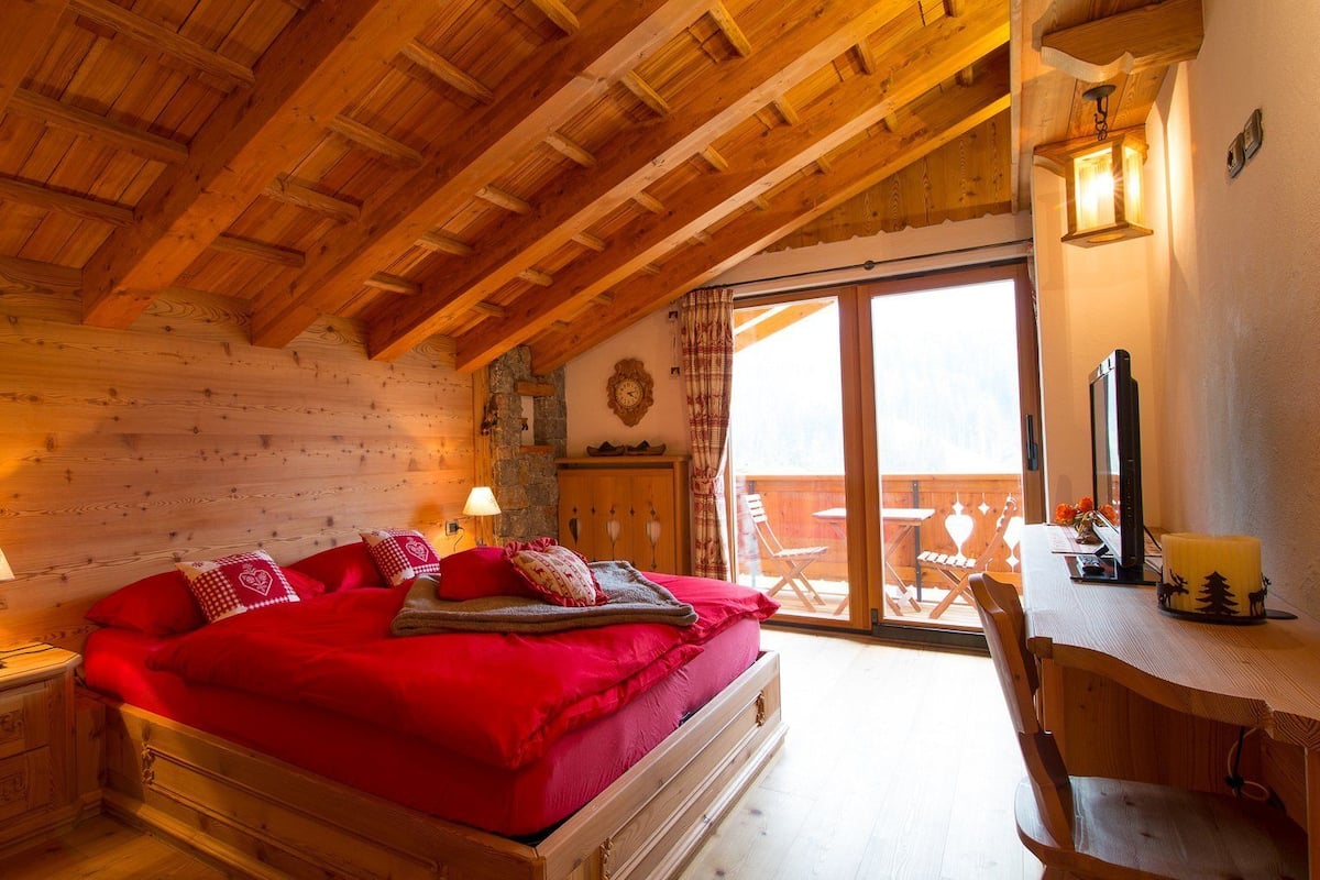 Un dormitorio acogedor con techo de madera, una cama grande con ropa de cama roja, un escritorio con una computadora y vistas al exterior.