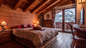 Un dormitorio rústico de madera con una cama grande, paredes de madera, un escritorio y vistas a árboles cubiertos de nieve.