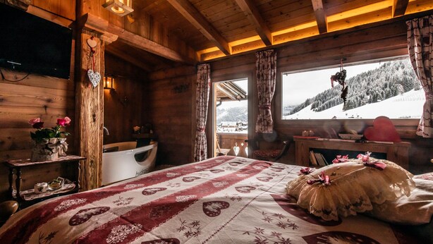 Cómodo cuarto de cabaña de madera con una cama, una ventana con vista a un paisaje nevado, un televisor de madera montado en la pared y una pequeña mesa con un jarrón de flores.