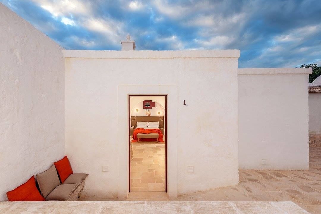 Une petite pièce aux murs blancs, avec un lit et une couverture rouge, une petite table avec une lampe, et une vue sur le ciel.