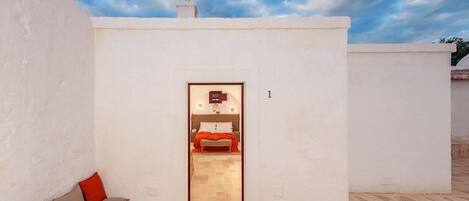 Une petite pièce aux murs blancs, avec un lit et une couverture rouge, une petite table avec une lampe, et une vue sur le ciel.