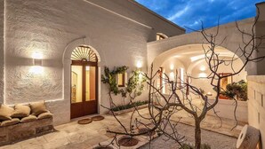 A courtyard with a stone bench, a tree, and a building with arched doorways and lighting. - Masseria Stefanodelconte (Fasano)