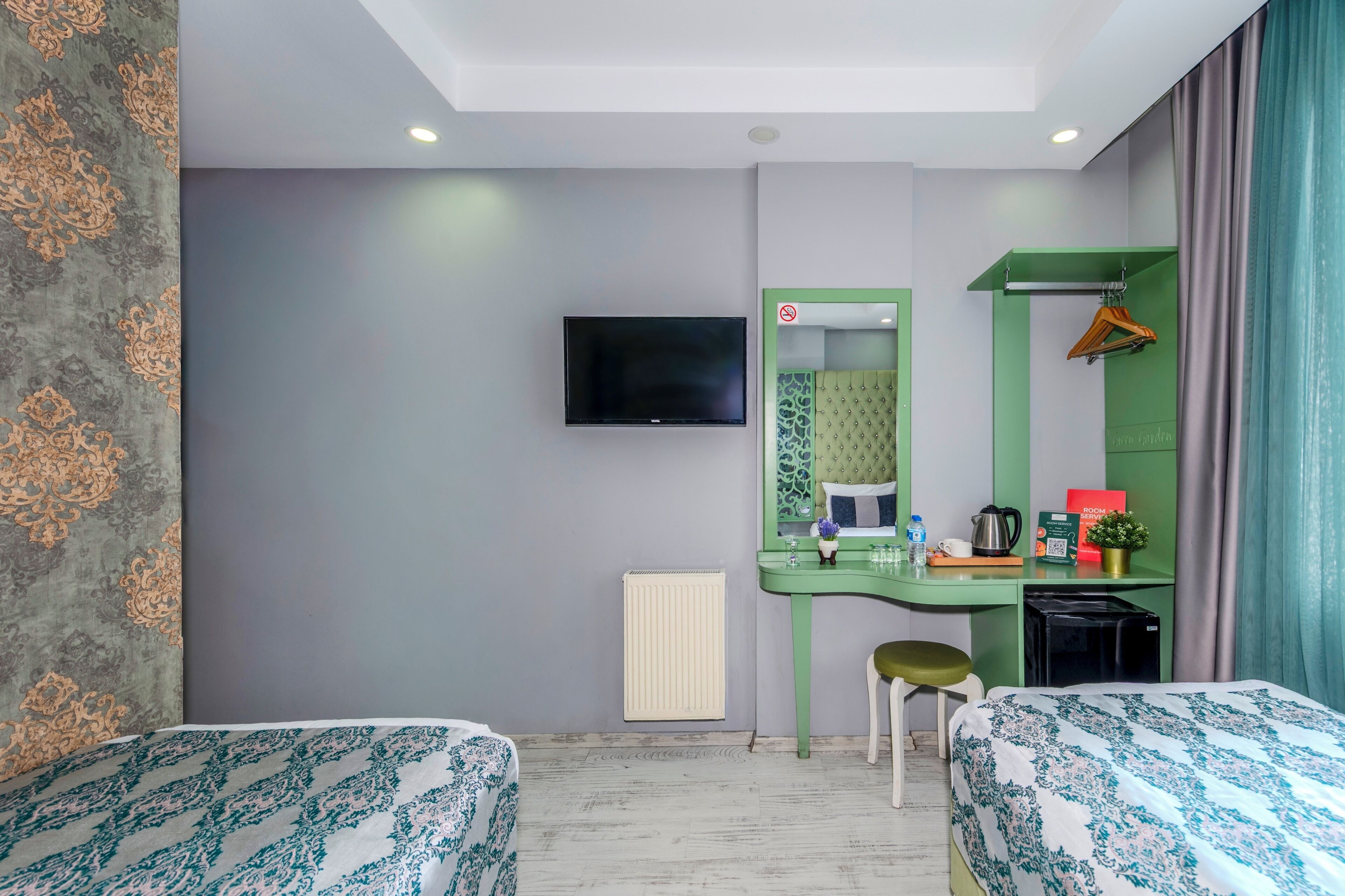Habitación de hotel con dos camas, un escritorio verde con espejo, un televisor montado en la pared, un radiador y una planta pequeña.