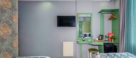 Habitación de hotel con dos camas, un escritorio verde con espejo, un televisor montado en la pared, un radiador y una planta pequeña.