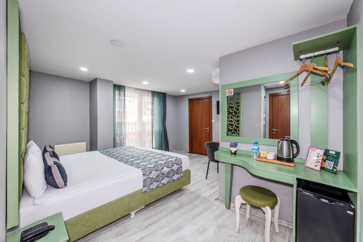 A hotel room with a bed, a desk, a chair, a small stool, a mirror, a radiator, and a door.