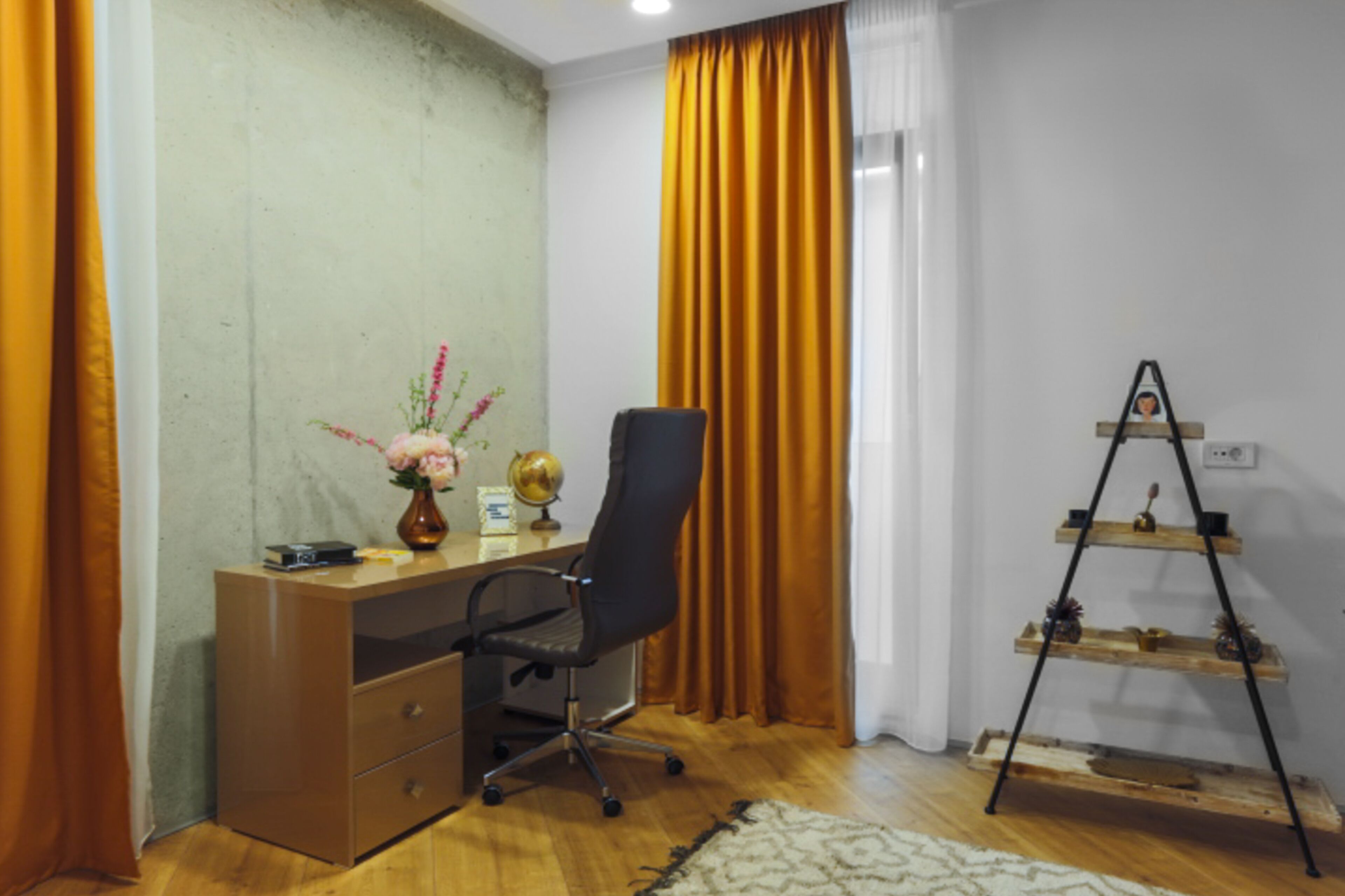A room with a wooden desk, a black office chair, a globe, a vase with flowers, and a wooden shelf with decorative items.
