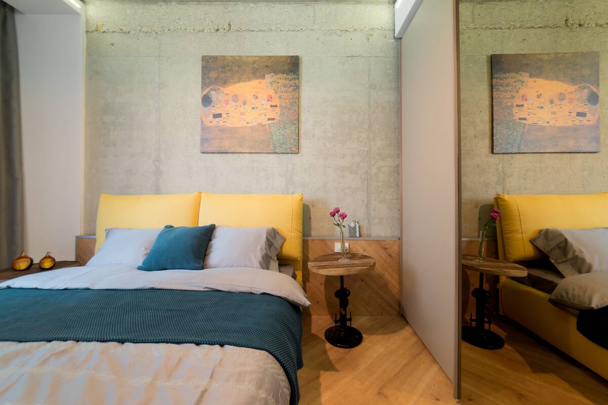 A bedroom with a bed, a small table with a vase, and two paintings on the wall.