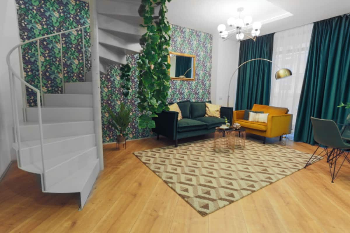 A modern living room with a staircase, a green sofa, a yellow armchair, and a patterned rug.