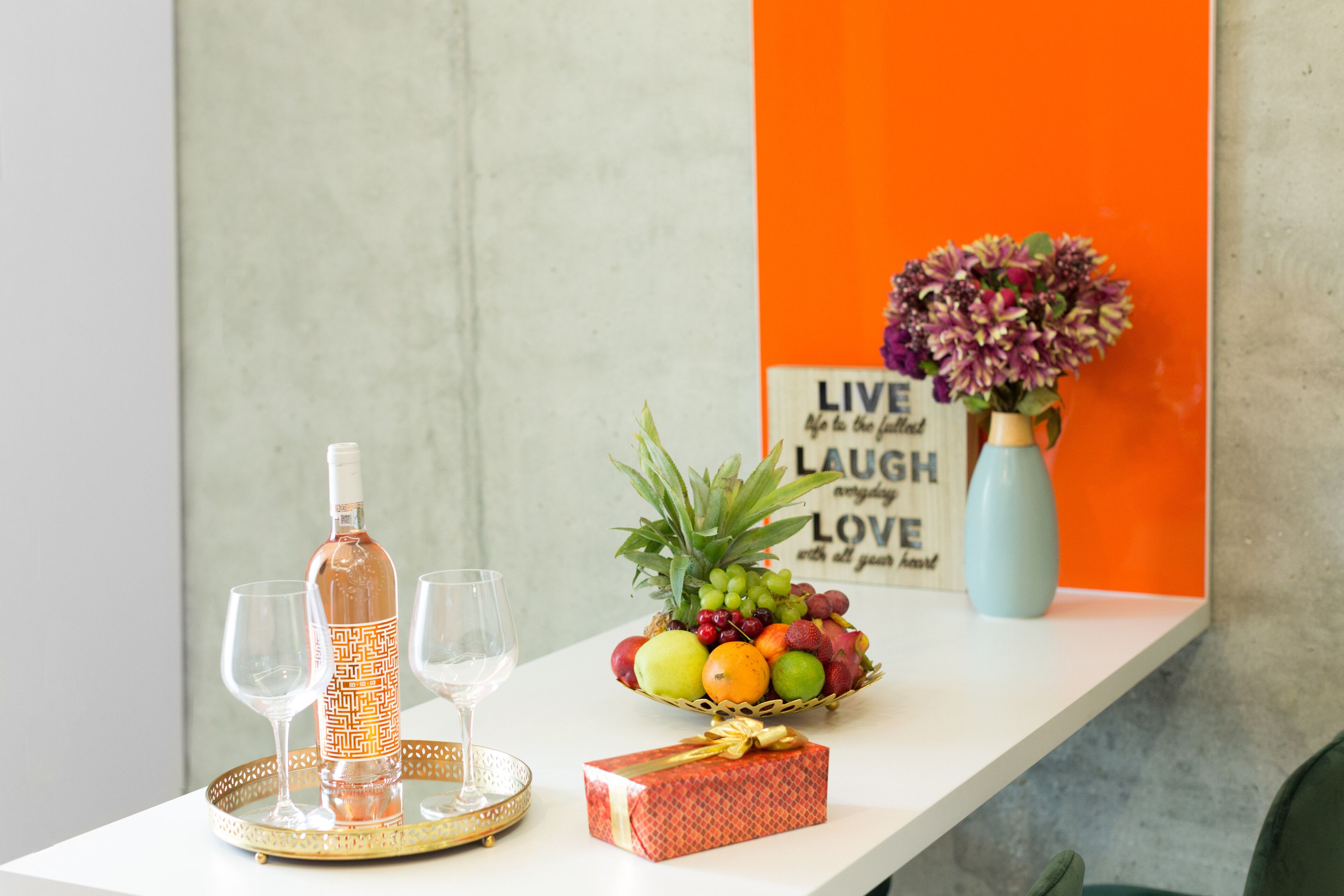 Una mesa con una botella, vasos, una cesta de frutas, un florero con flores y una caja de regalo.