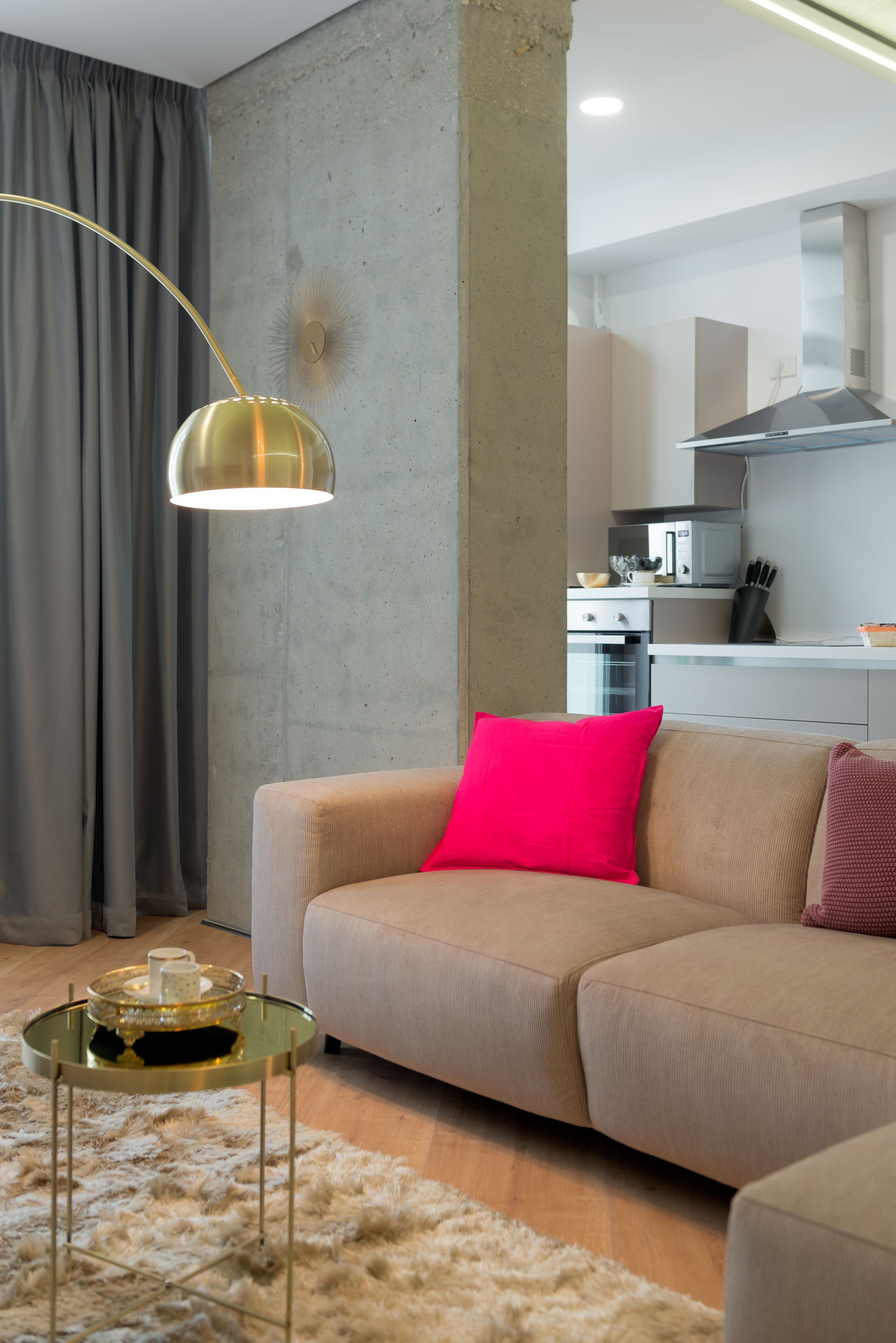 Una sala de estar con un sofá color beige, una mesa de centro de vidrio, una lámpara colgante dorada y una cocina al fondo.