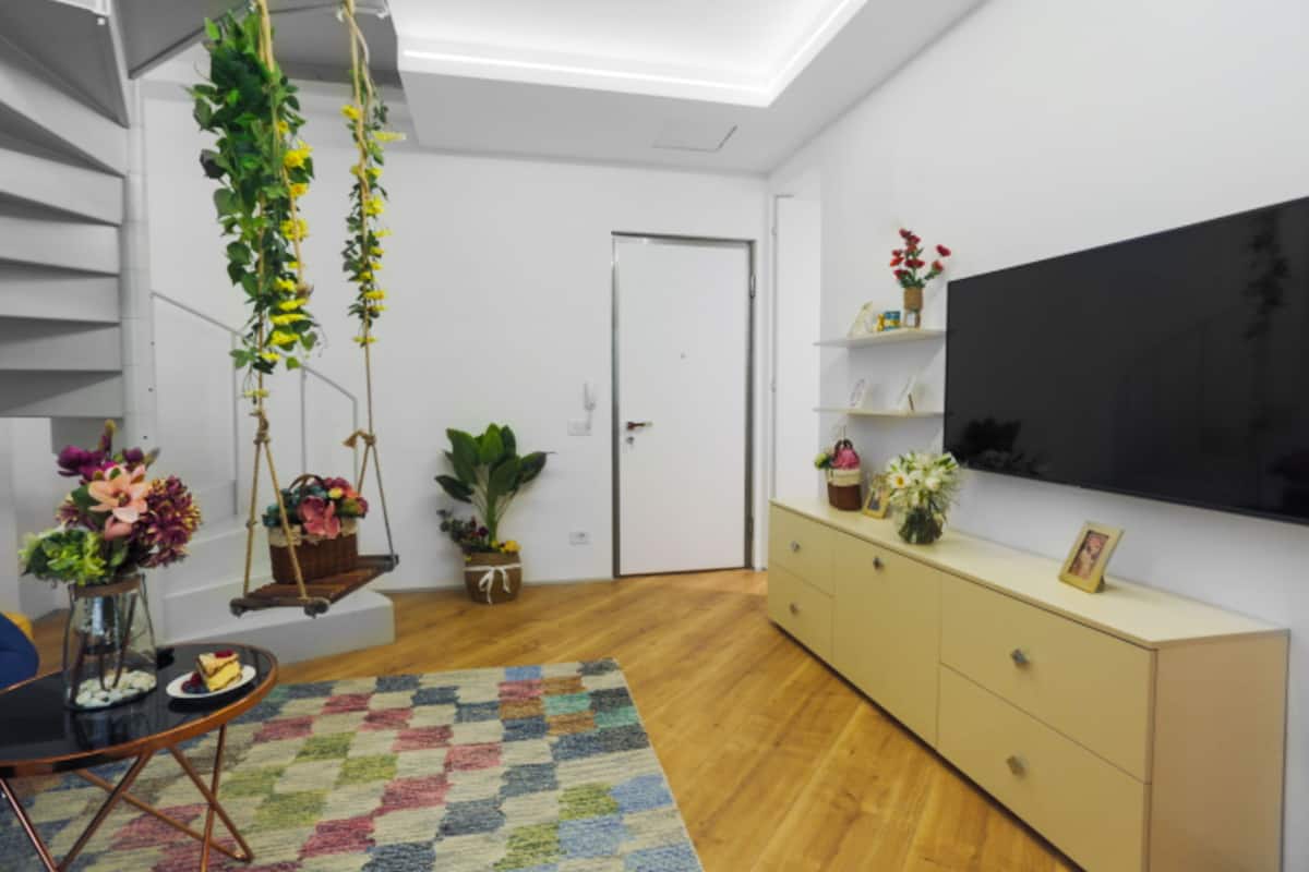 A living room with a black TV, wooden flooring, a colorful rug, a sofa, a dining table with a cake, a hanging plant, and a cabinet with decorative items.