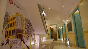 Um corredor bem iluminado com um mural de um edifício e pessoas, uma escadaria e portas cor de verde-água.