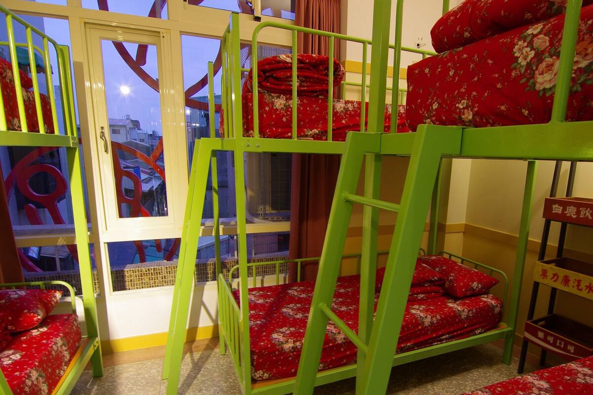 Beliches com roupa de cama florida, uma janela com cortinas e vista para prédios.