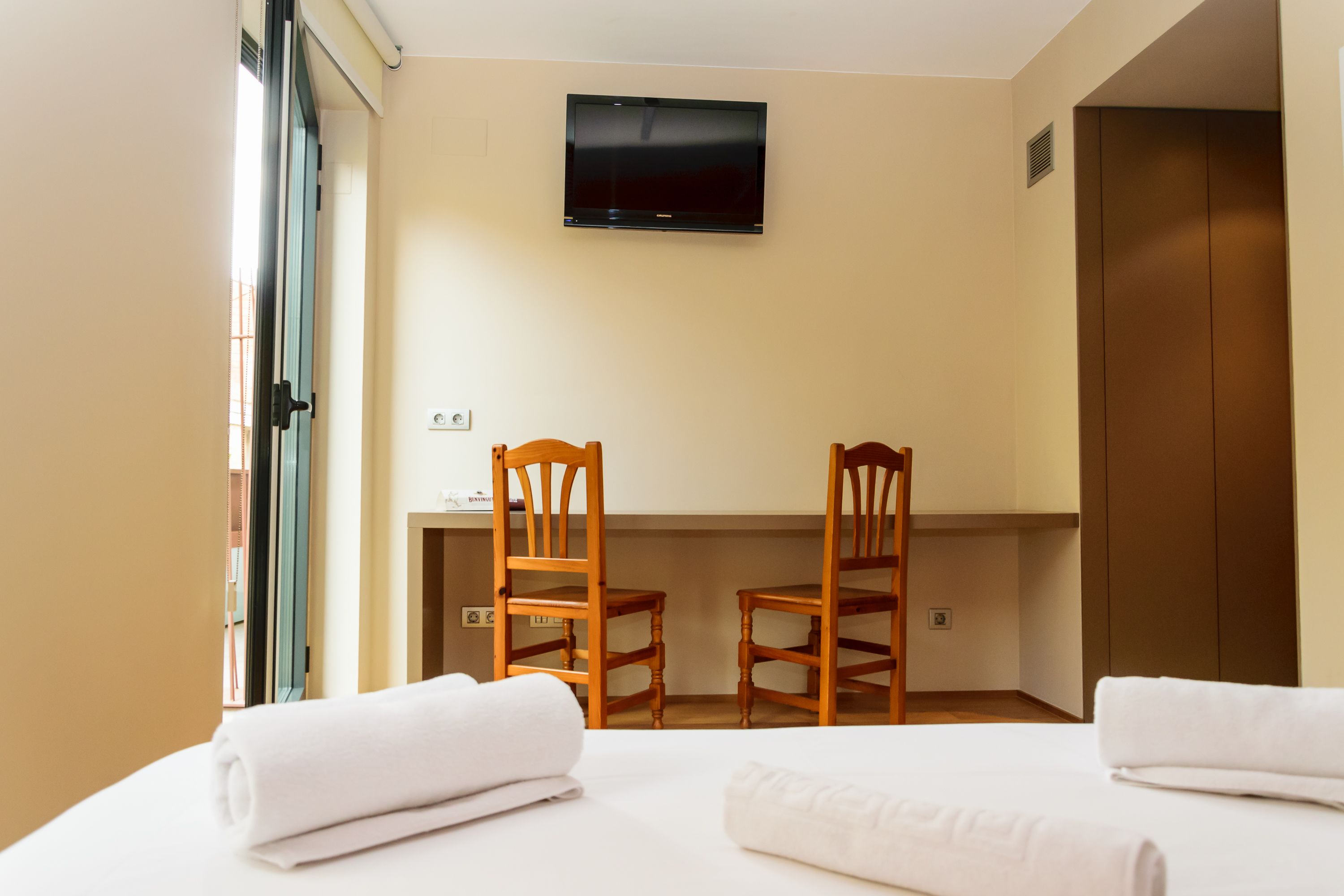 Habitación de hotel con una cama, dos sillas de madera, un televisor de pantalla plana montado en la pared y una puerta.