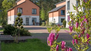 A residential area with two houses, one with a white and orange facade and the other with a white and grey facade. There is a well-maintained lawn and a tree with pink flowers in the foreground. - Ferienpark Rübezahl am Müggelsee (Berlin)