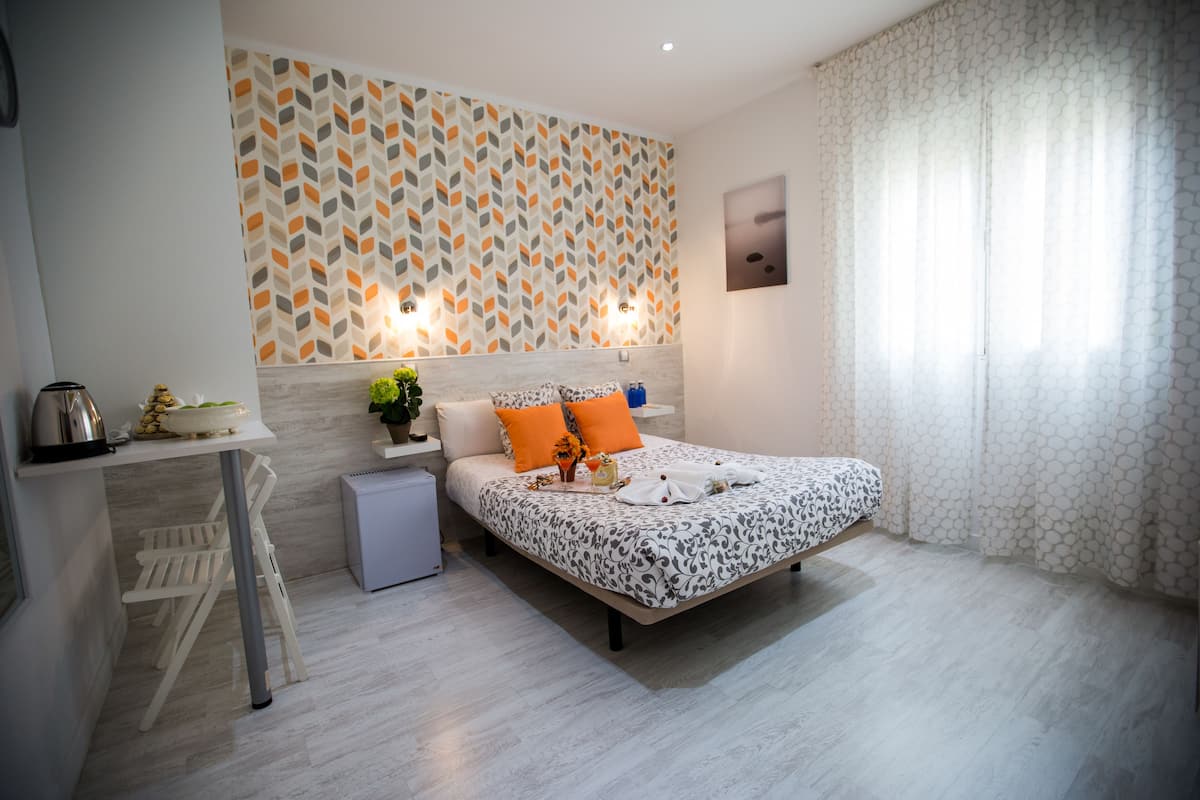 A modern bedroom with a patterned wall, a bed with orange pillows, a bedside table with a kettle, and a small bedside plant.