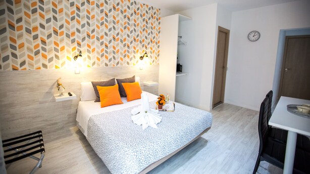 Una habitación de hotel moderna con papel tapiz estampado, una cama bien tendida con almohadas naranjas, una mesita de noche con lámpara y una pequeña mesa con un jarrón de flores.