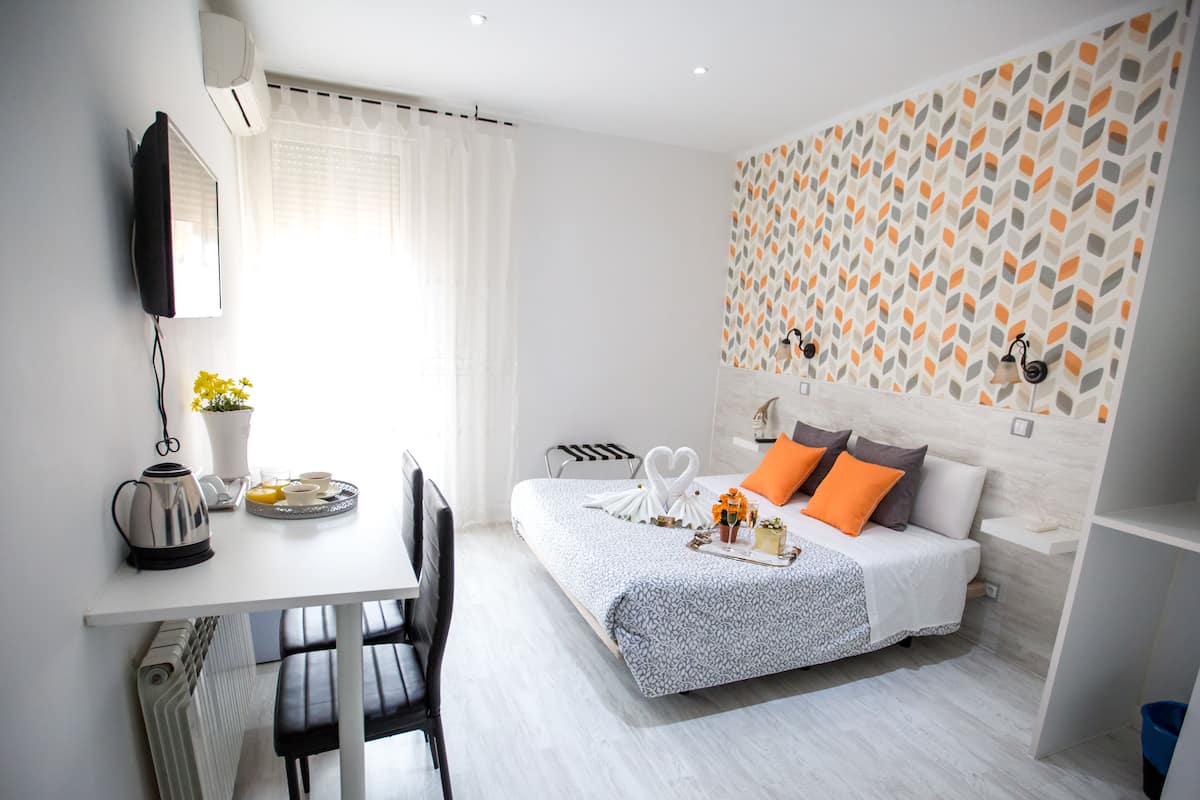 A modern hotel room with a bed, a desk, a chair, a radiator, a potted plant, and a wall with a geometric pattern.