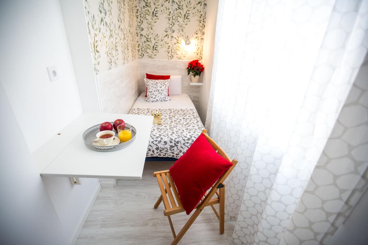 A small, neatly arranged bedroom with a single bed, a bedside table with a tray of fruit, and a chair with a red cushion.