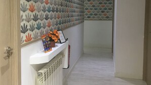 Un pasillo con papel tapiz estampado, un radiador blanco y un estante pequeño con un cuadro enmarcado y flores.