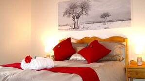 A neatly made bed with red pillows, a white towel with a red flower, and a painting of trees on the wall. - Sunset Cottage- Noordhoek (Cape Town)