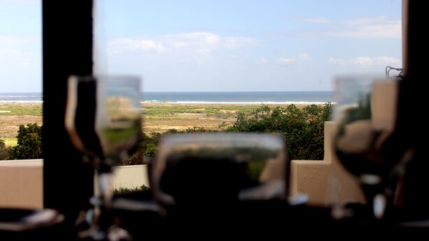 A view from a window with two chairs and a table, overlooking a landscape with a body of water and greenery. - Sunset Cottage- Noordhoek (Cape Town)