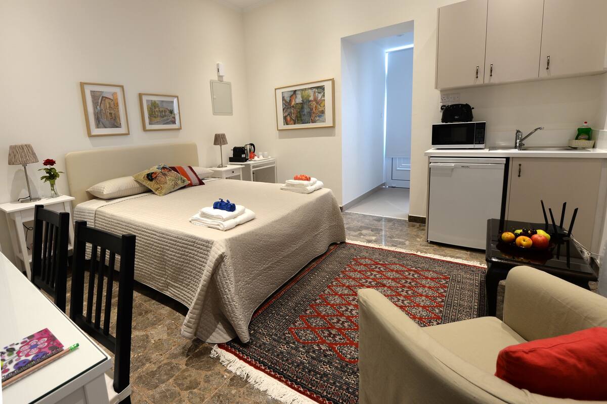 A hotel room with a bed, a desk, a chair, a small kitchen area, and a bathroom visible through an open door.