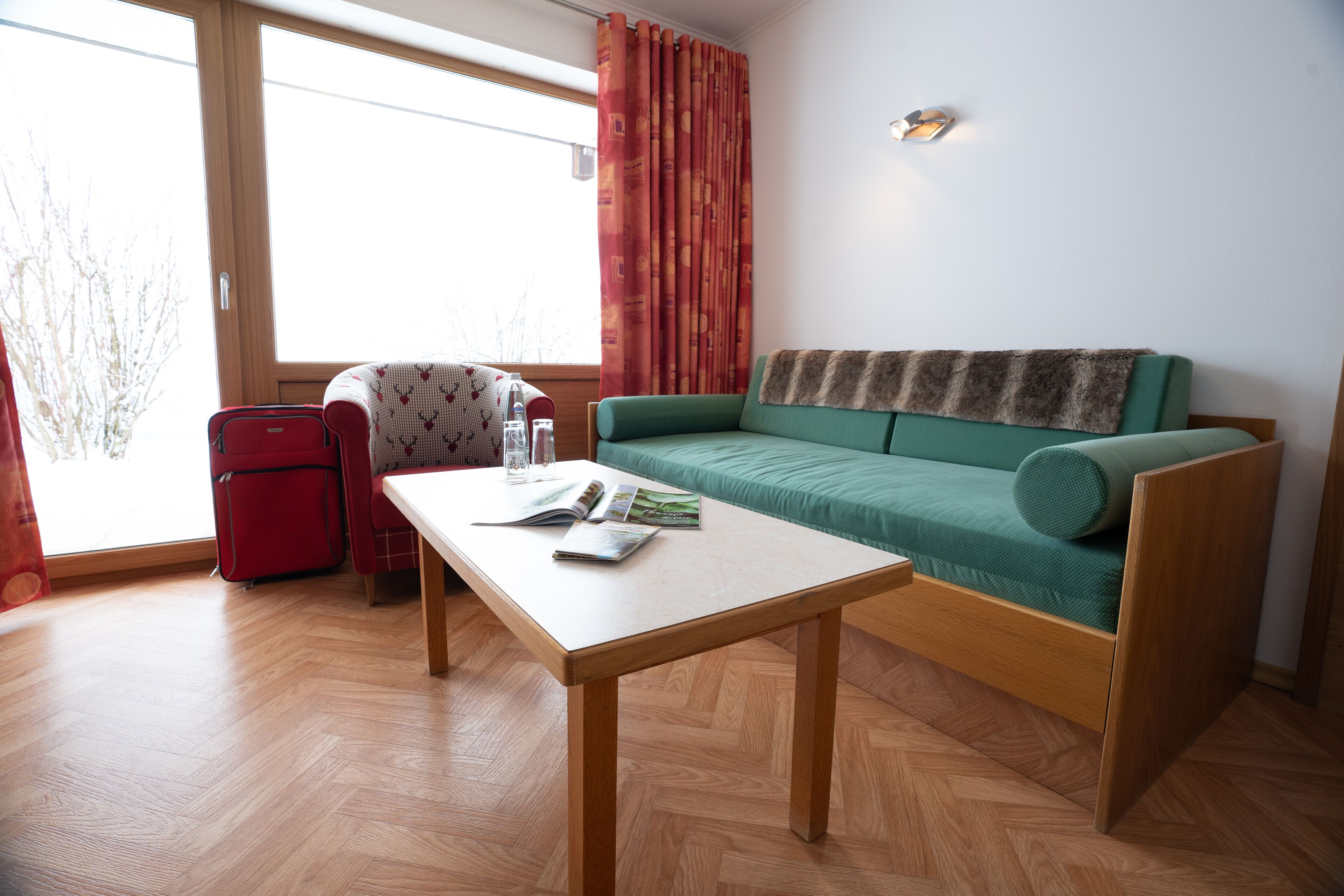 A room with a wooden floor, a green sofa with a fur throw, a red suitcase, and a table with magazines.