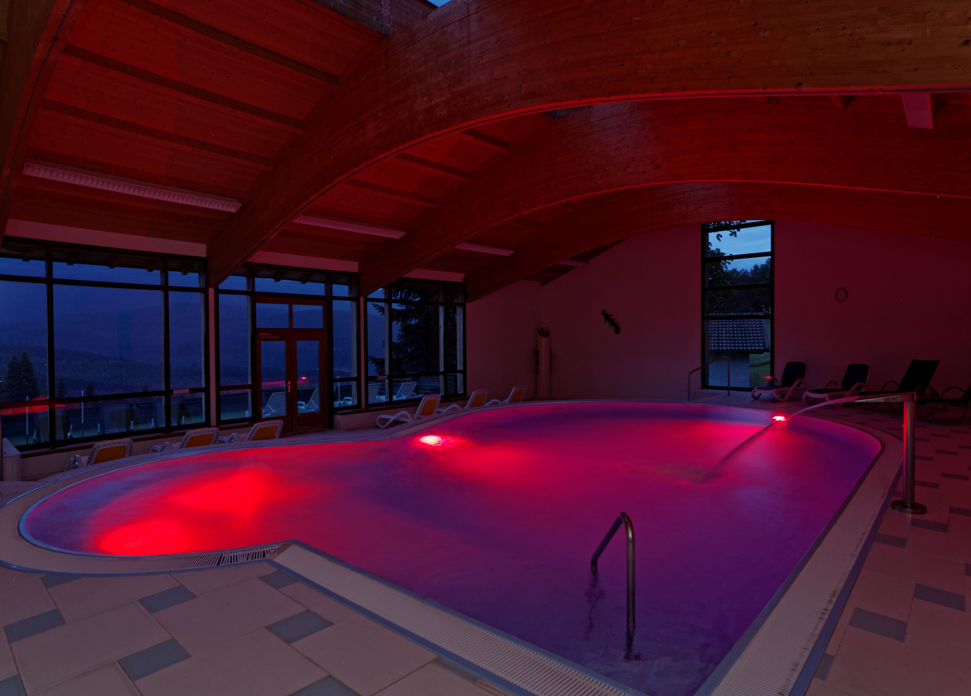 An indoor swimming pool with a red-lit edge, lounge chairs, and large windows offering a view of the outside.