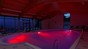 An indoor swimming pool with a red-lit edge, lounge chairs, and large windows offering a view of the outside.