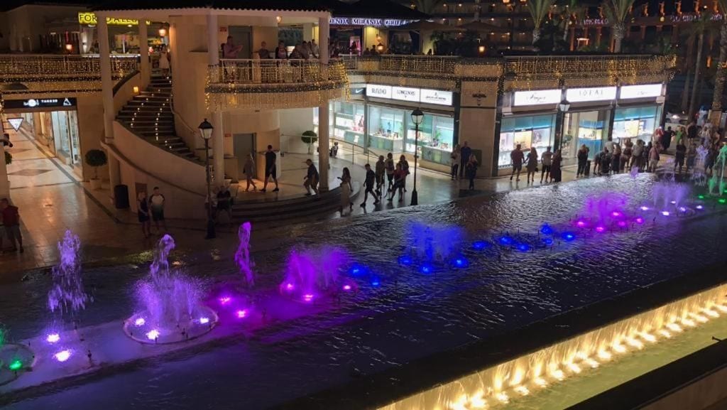 Ett offentligt torg med fontäner och ett vattenspel, omgivet av butiker och en trappa.