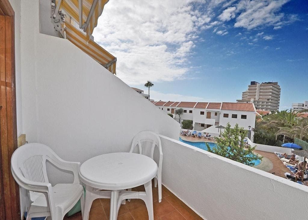 A balcony with two white plastic chairs and a small round table, overlooking a swimming pool and buildings in the background.