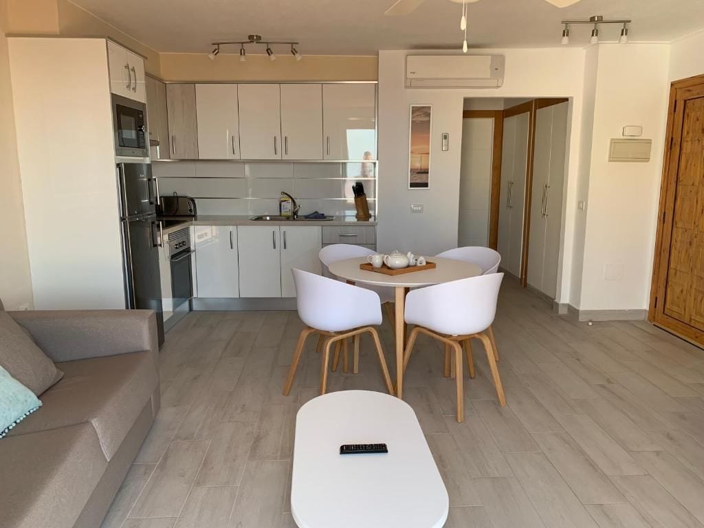 Una cocina moderna con armarios blancos, una mesa de comedor con sillas, un sofá y un control remoto en el suelo.