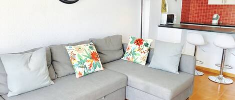 A modern living room with a grey sectional sofa, a round wall clock, a floor lamp, and a kitchen area with a red backsplash.