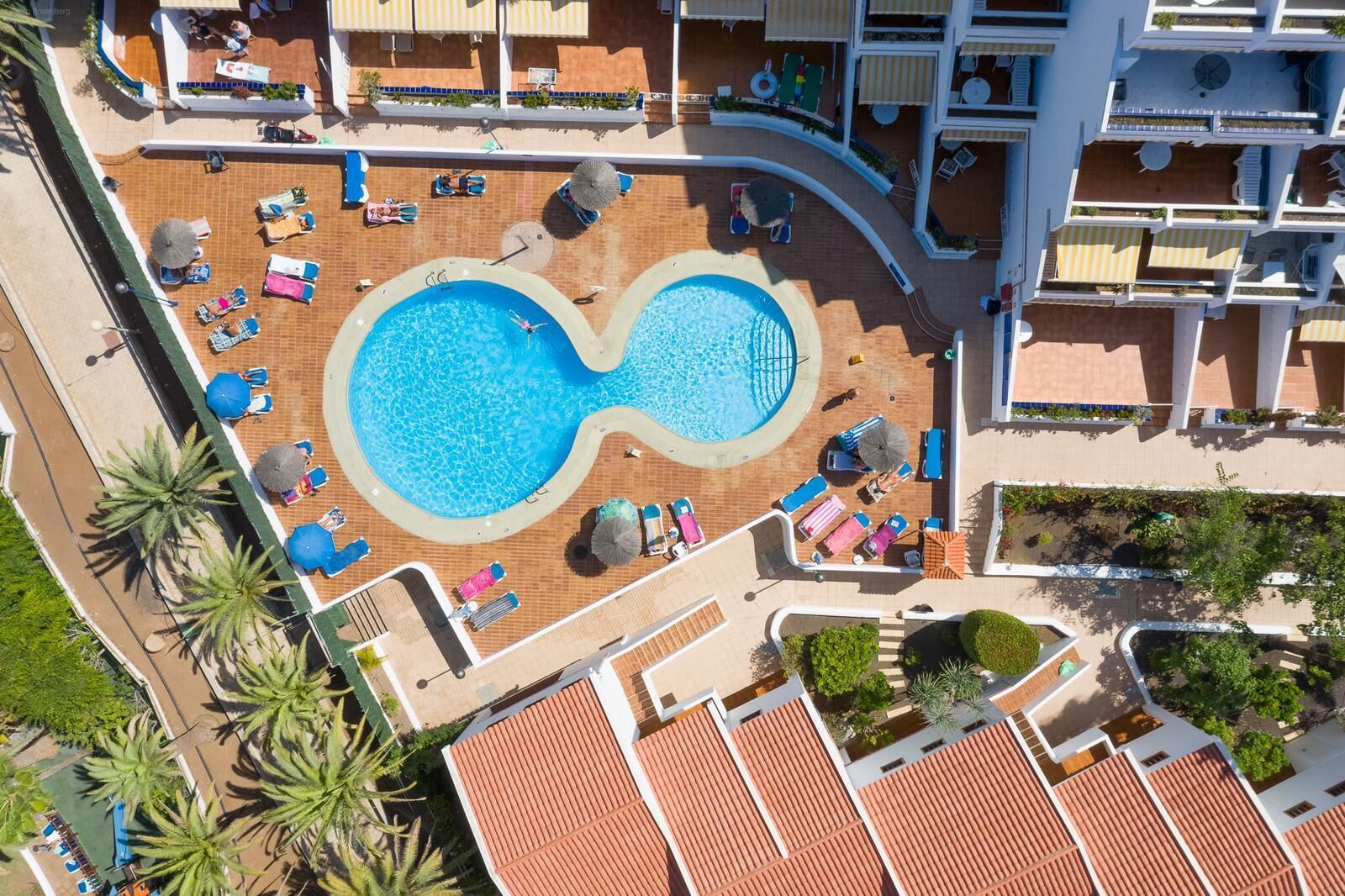 An infinity pool surrounded by lounge chairs and umbrellas, with a building complex in the background.
