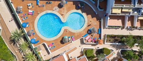 An infinity pool surrounded by lounge chairs and umbrellas, with a building complex in the background.