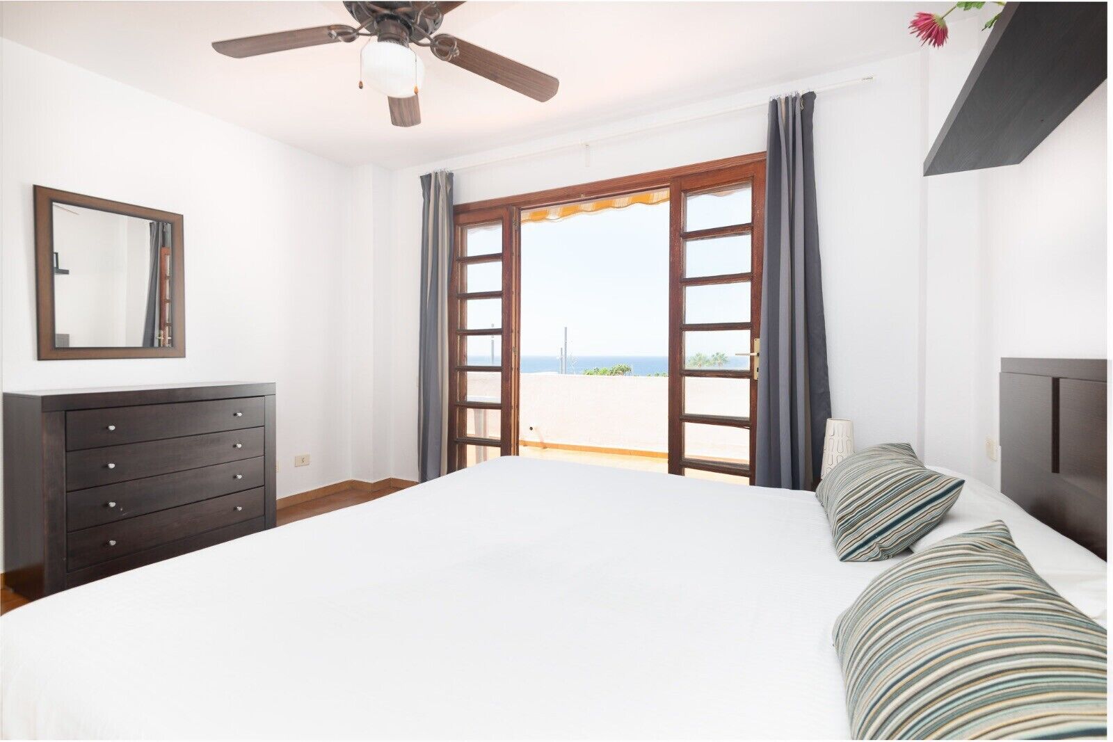 Un dormitorio con una cama grande, un aparador, un ventilador de techo y vistas al mar a través de un ventanal amplio.